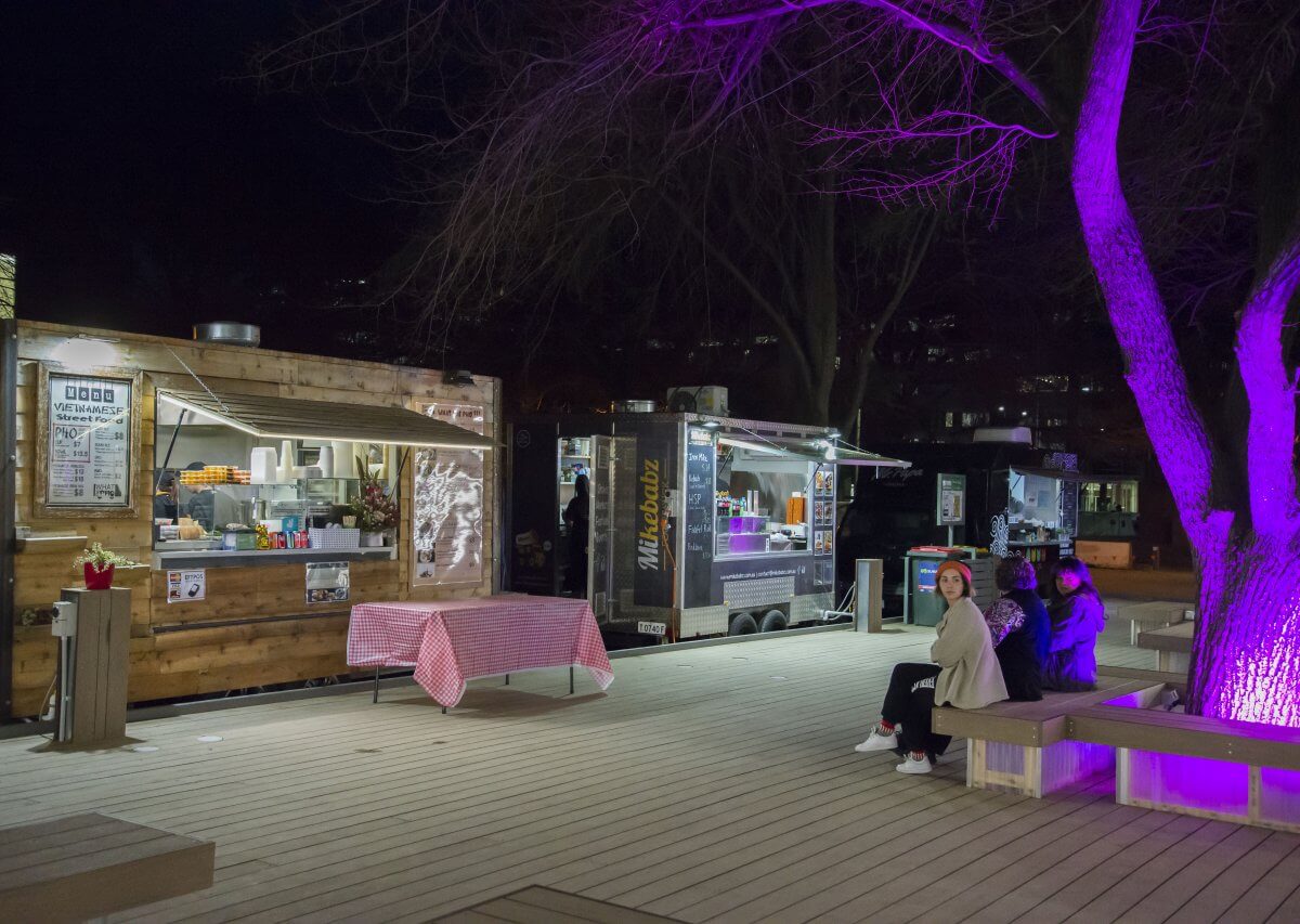 The Pop-Up deck at night with two food stalls, and an illuminated purple tree.
