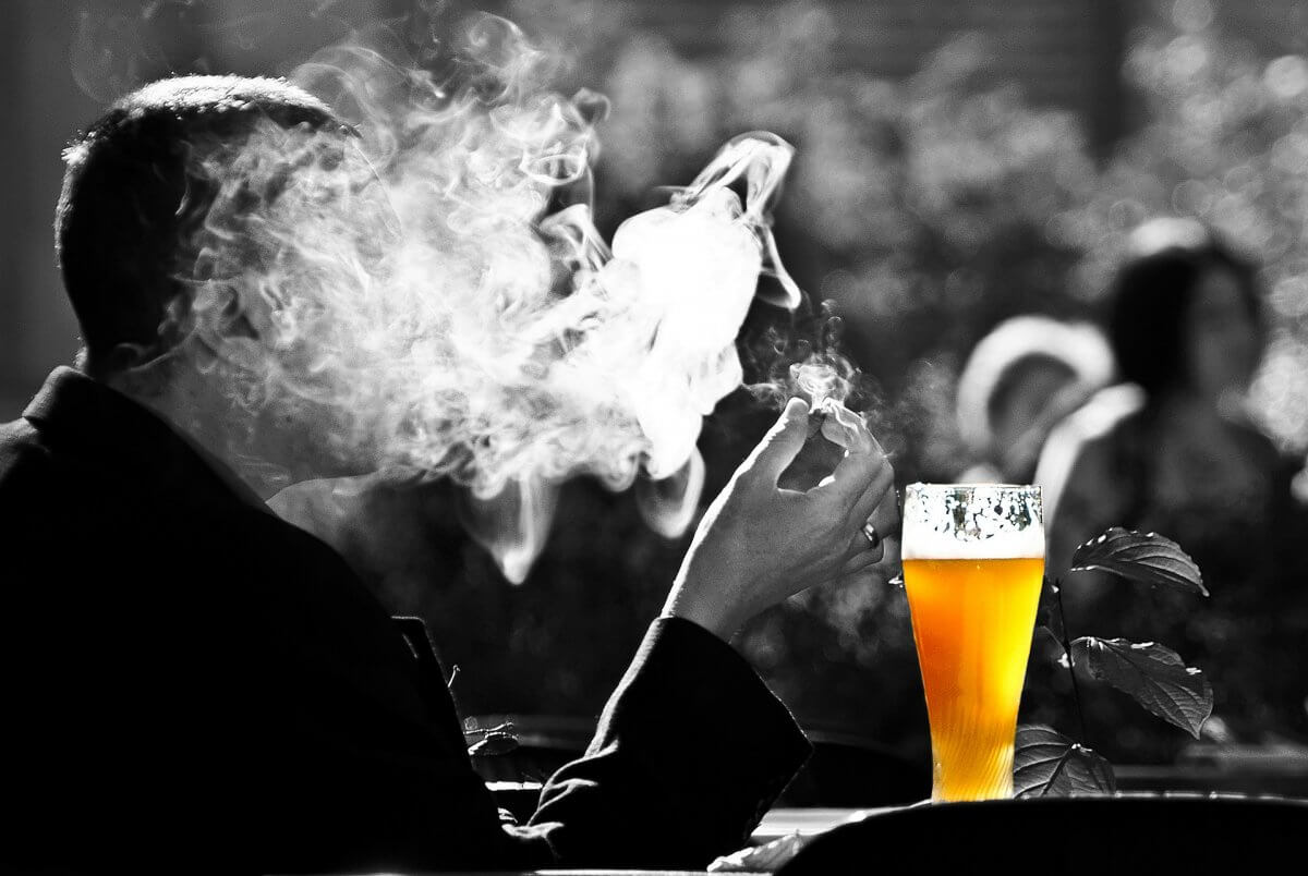 Male figure smoking a cigarette. A beer is placed beside him on the table.