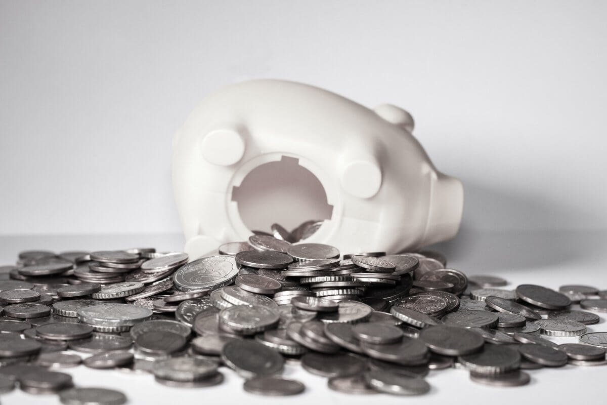 A white piggy bank on its side, with many silver coins spread out in front of it.