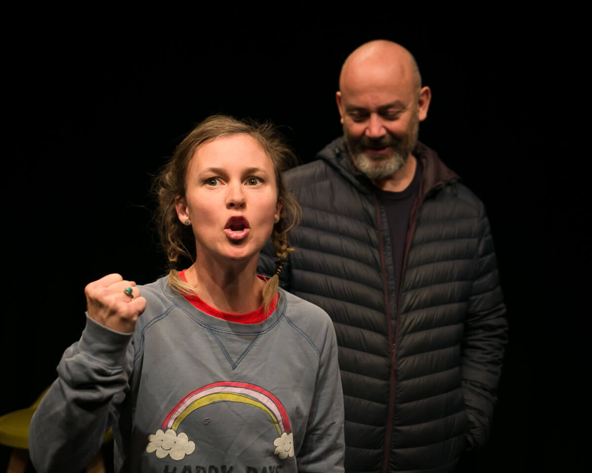 Beth Buchanan stands facing camera with her fist clenched in mid-speech. She is wearing a jumper with a rainbow. Adriano Cortese, wearing a puffy black jacket, stands behind her staring down.