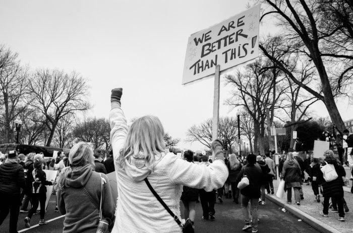 Black and white image of a protest. A woman is holding a sign which reads, ' We are better than this'