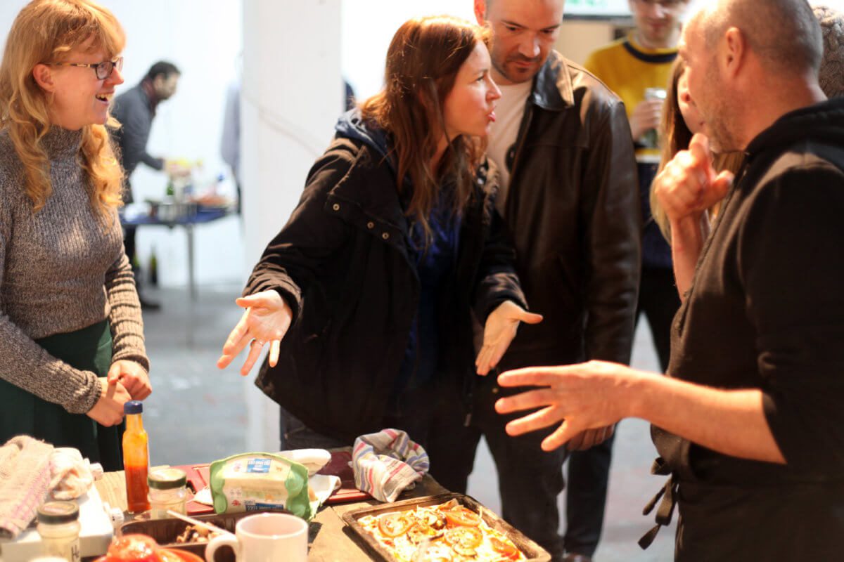 A group of people discussing with each other what to put on their pizza. A woman centre is indicating to the pizza with passion.