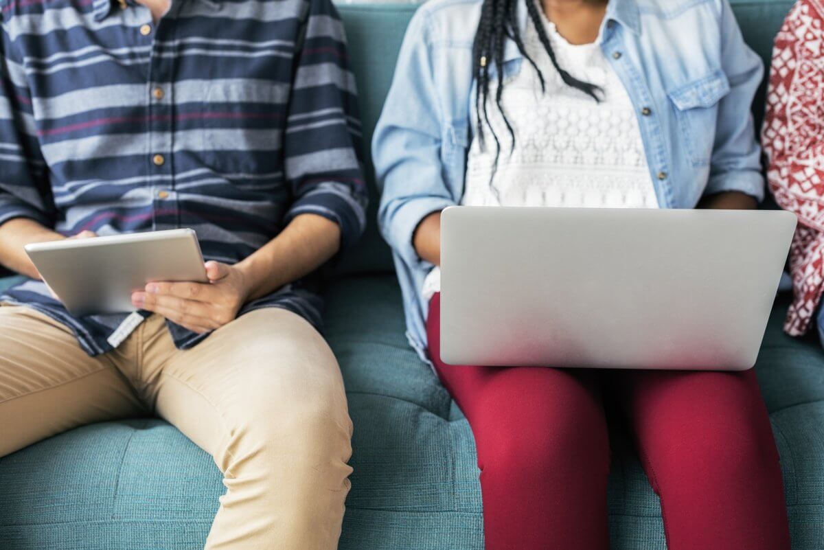 image shows two students, both with laptops. the one on the left is wearing a stripey blue shirt with cream pants while the student on the right is wearing a blue cardigan with red pants