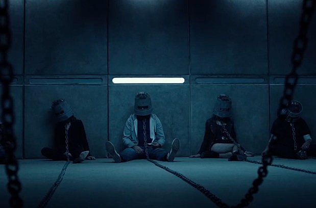 Still from the film, featuring 4 people chained up and wearing heavy metal masks over casual clothes in a concrete room with only a single slit window