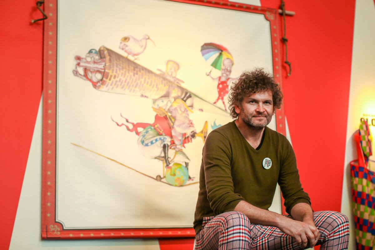 David Rowe, wearing a green jumper and red tartan pants, sits in front of the exhibition's hero cartoon, which he drew. The cartoon shows a circus scene. Various political figures, such as Donald Trump and Malcolm Turnbull, are depicted as performers.