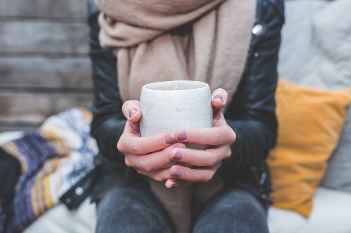 A person holding a large cup of tea in their two hands. Person is wearing a creme scarf and black jacket