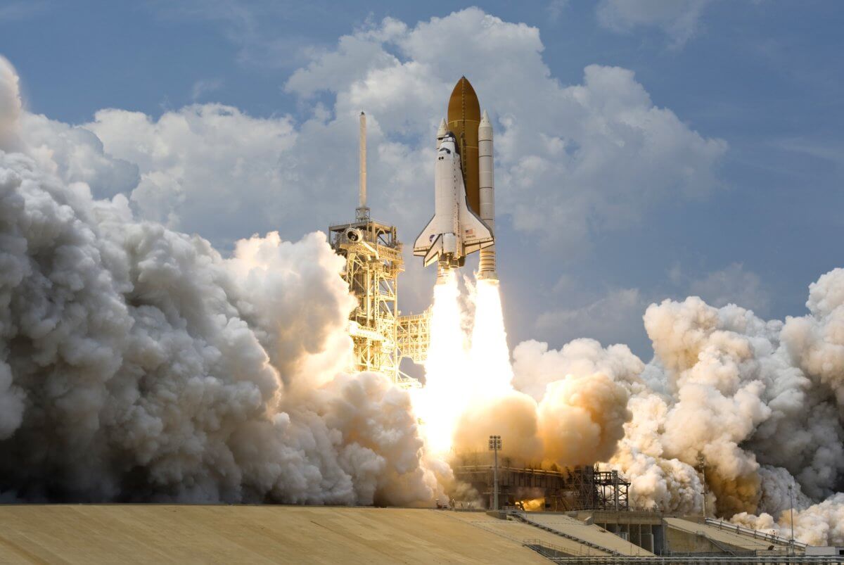 Photograph of a rocket launching directly up. Clouds of smoke surround the launch pad.