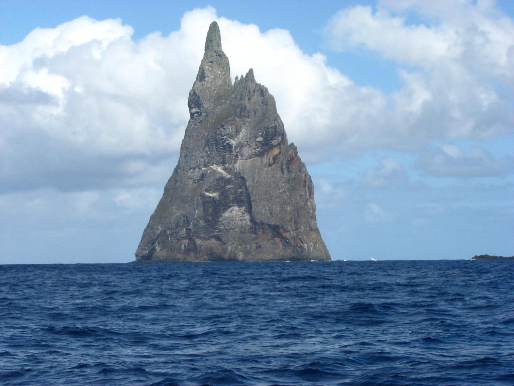 Photograph of Ball's Pyramid. The picture features a rocky island in the middle of the photo, with the sea on the bottom half of the image, and a cloudy sky in the top half.