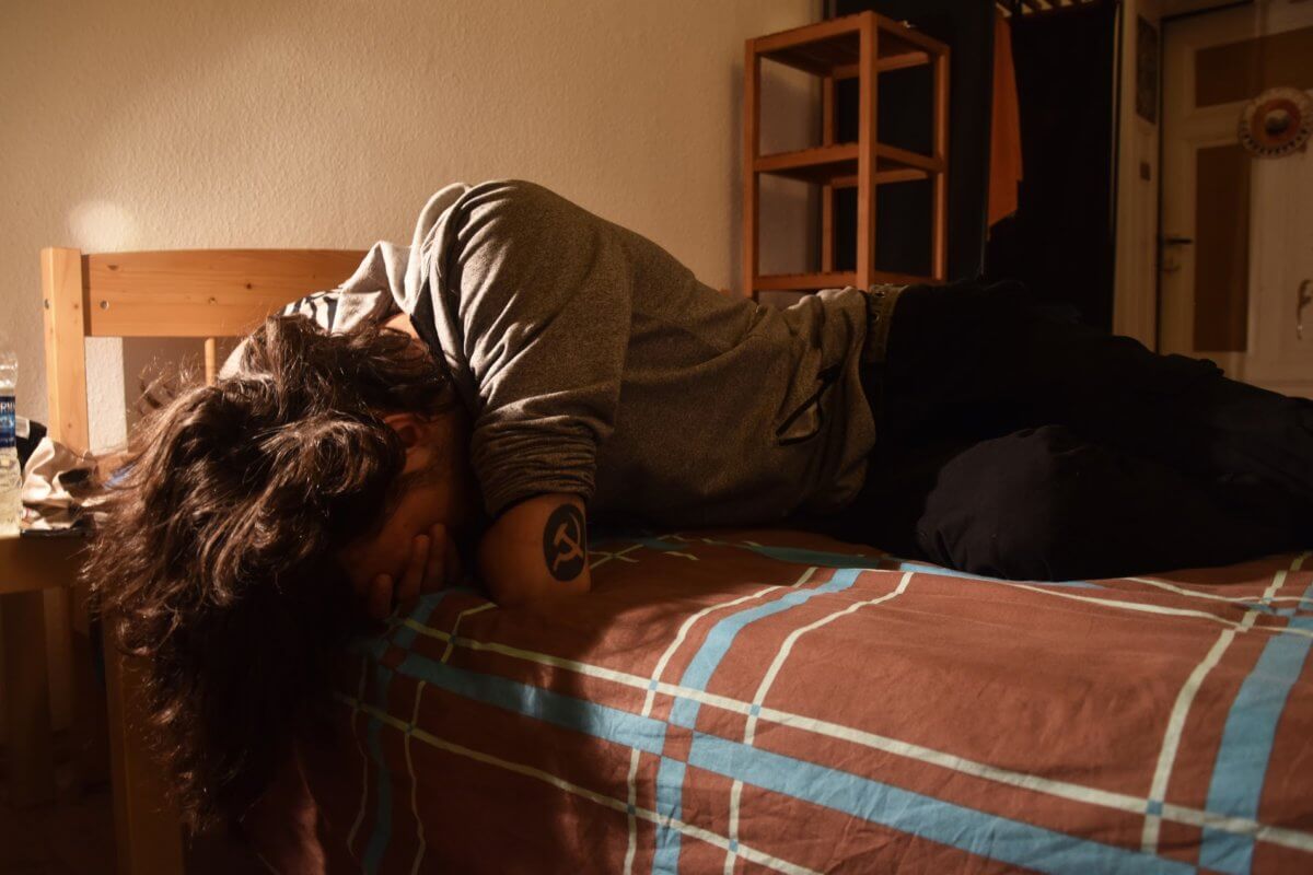 A young man hides his face with his arm while lying on a bed.