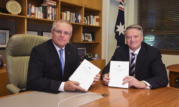 Scott Morrison and Matthias Corman seated together in an office, holding copies of the MYEFO. An Australian Flag is prominent behind them.