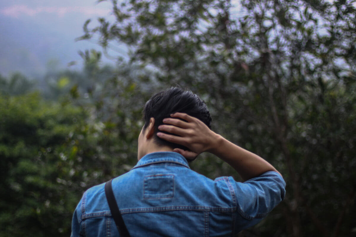 A man stands in the center of the photo with his back to the audience - and his right hand placed on the back of his head. He is facing green bushes and trees, which fill up the remainder of the photo.