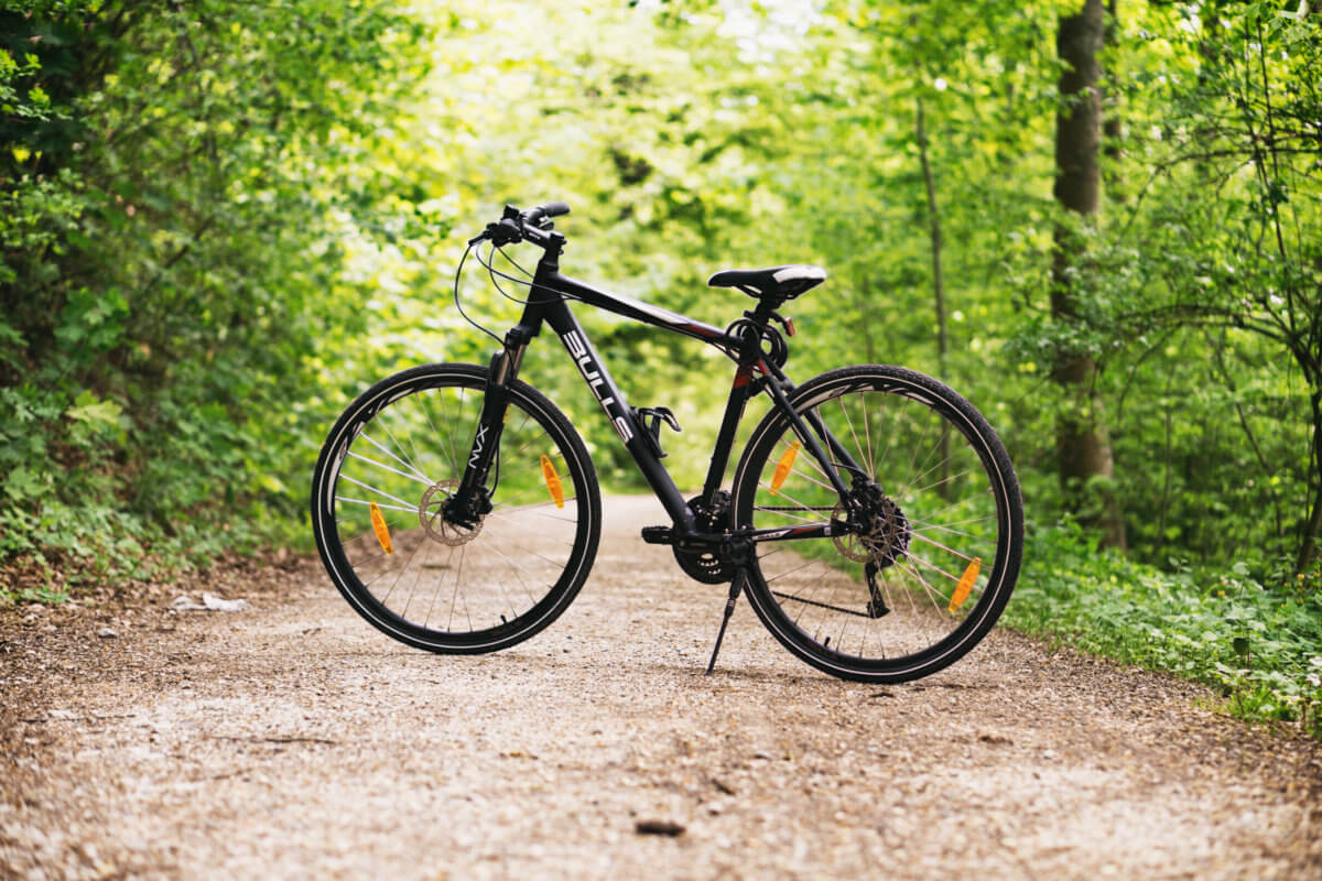 Image has a bicycle in the front and focus of the picture. The bike is on a dirt track, and there is a forest background behind the bike.