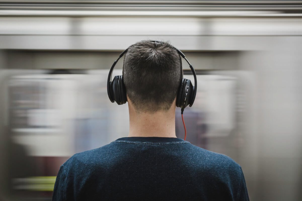 A man stands on a platform before a blurred, speeding train. He has short black hair, is wearing a casual navy jumper and - most importantly - headphones.
