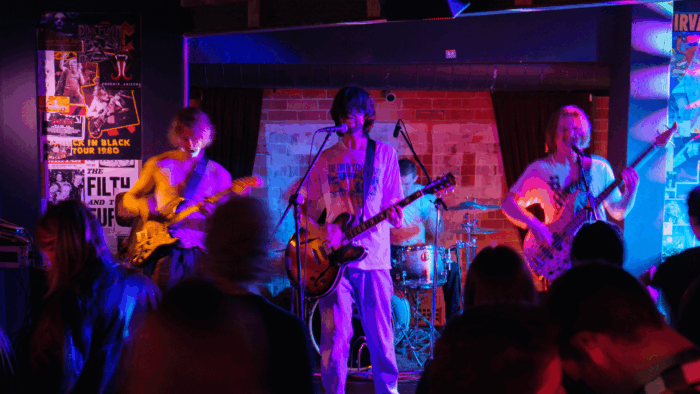 four rock musicians play to an intimate crowd under ambient lights and ripped band posters